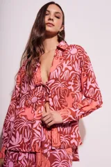 Blusa de poliéster con estampado de flores y animales en tonos rosa y marrón. Tiene cuello camisero, mangas largas y cierre frontal con lazo.