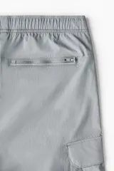 Pantalón cargo de nylon color gris claro, con cintura elástica y cordón ajustable. Presenta bolsillos laterales con cierre, bolsillos cargo con solapa en las piernas y cordón ajustable en los bajos.
