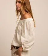 Blusa de tricolina blanca con estampado de lunares dorados en foil, escote descubierto estilo hombro a hombro y mangas largas abullonadas con puños elásticos.