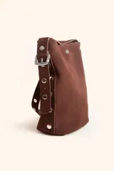 Cartera tipo balde color tostado, confeccionada en gamuza, con correa ajustable decorada con tachas metálicas plateadas.