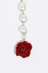 Collar largo tipo lazo compuesto por perlas irregulares de vidrio, con cierre frontal de barra y argolla en tono dorado. Finaliza en una sección colgante que termina con un dije en forma de rosa roja de resina.