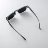 Lentes de sol polarizados con armazón gris translúcido y diseño rectangular, con detalle triangular negro en las patillas y cristales oscuros.