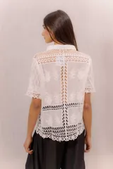 Camisa de algodón color crudo, con cuello camisero, mangas cortas y cierre frontal con botones. Presenta paneles de tejido calado y detalles bordados transparentes.