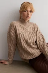 Polerón de punto color beige con cuello alto y diseño de trenzas y texturas en relieve. Presenta mangas largas con puños acanalados y un corte holgado.