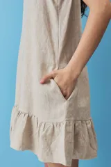 Vestido corto de lino color beige, de silueta suelta y sin mangas, con escote en V decorado con un botón, bolsillos laterales y detalle de volado en el ruedo.