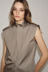 Camisa sin mangas confeccionada en algodón, con cuello redondo y detalle de lazada frontal. Presenta cierre frontal con botones ocultos por solapa y bajo redondeado.