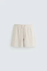Bermuda de corte relajado color beige claro, confeccionada en lino. Presenta cintura elástica con cordón ajustable y bolsillos laterales y traseros.