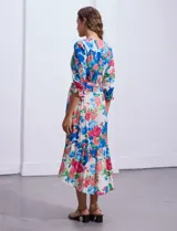 Vestido midi con estampado floral en tonos azules, rojos y blancos. Tiene escote en V, mangas 3/4 con puños fruncidos, cintura elastizada con lazo de ajuste y falda con volantes.
