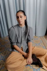 Chaqueta gris de silueta femenina con mangas abullonadas, cuello redondo y cierre frontal con botones forrados. Presenta detalles de bordado artesanal a mano en los hombros y solapas.