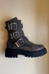 Bota de caña media confeccionada en cuero color gris oscuro con efecto desgastado. Presenta tres correas laterales con hebillas metálicas grandes y una suela track negra de plataforma dentada.