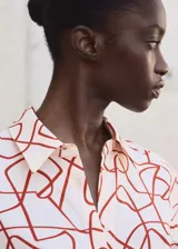 Camisa de corte recto con estampado abstracto en tonos rojizos sobre fondo claro. Presenta cuello camisero, cierre frontal con botones ocultos bajo tapeta y mangas largas con puños abotonados.