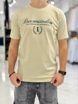 Remera color beige con cuello redondo y mangas cortas. Estampado frontal con el texto "Duxonstudios" en tipografía cursiva, acompañado de un logotipo circular con hojas de laurel y una figura central en tono verde oscuro.