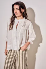 Blusa de algodón peinado color off-white, con cuello mao, cierre frontal de botones, mangas tres cuartos abullonadas con puños con volados, detalles de alforzas y apliques de encaje en el frente y los hombros.