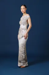 Vestido largo de corte ajustado con escote asimétrico tipo bote que deja un hombro al descubierto. Presenta un estampado artístico de paisaje en tonos neutros, tierra y gris sobre fondo claro, con un sutil fruncido en la zona de la cintura.