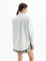 Camisa de poplin color crema, de corte clásico con cuello camisero, mangas largas y cierre frontal con botones dorados de media esfera.