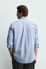 Camisa de corte regular fit confeccionada en tejido de algodón, con diseño de rayas verticales en tonos azules. Presenta cuello abotonado, manga larga con puños abotonados, cierre frontal de botones y un bolsillo en el pecho.