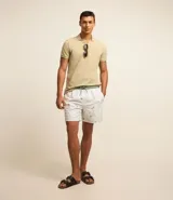 Bermuda de baño para hombre confeccionada en microfibra ligera, color blanco, con una estampa sutil de cocoteros en tonos verdes/azulados. Presenta cintura elástica con cordón de ajuste contrastante en color verde, y bolsillos laterales.