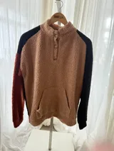 Buzo de corderito con diseño tricolor, que combina cuerpo en tono tostado, una manga negra y otra en color ladrillo. Cuenta con cuello alto con botones frontales y bolsillo tipo canguro.