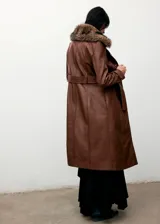 Abrigo largo de cuero sintético en color marrón, con diseño recto, cuello de solapa con pelo sintético extraíble, cierre frontal con botones y cinturón ajustable con hebilla. Cuenta con bolsillos laterales y mangas largas.