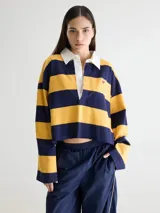 Remera estilo rugby oversize de manga larga y largo cropped, con diseño a rayas horizontales en verde oscuro y rosado. Posee cuello camisero blanco en contraste, cartera de botones, pequeño bordado en forma de corazón en el pecho y terminación desflecada en el ruedo.