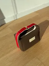 Necessaire de neopreno color marrón con manija superior en color rojo y detalles en crema. Cuenta con cierre superior y placa frontal con el logo de la marca.