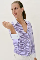 Camisa de popelín con diseño de rayas verticales en tonos blanco y lila, cuello solapa con escote en pico, manga larga y puños anchos contrastantes en color blanco. Cierre frontal con botones metálicos dorados.