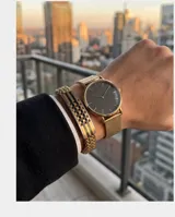 Pulsera de acero con baño de oro, compuesta por una cadena de eslabones trenzados y una placa central rígida con diseño geométrico de cuadros en tonos dorados y negros.