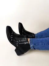 Botín corto de cuero negro con textura combinada en la puntera y el talón. Cuenta con punta fina, apliques metálicos en forma de tachas y cierre lateral plateado.