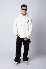Buzo tipo canguro de corte oversize, color blanco, con capucha y cordones ajustables. Presenta un pequeño estampado de una rosa roja en el pecho y un bolsillo frontal tipo canguro.