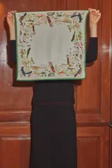 Pañuelo cuadrado de seda con estampado botánico y de fauna, que incluye ilustraciones de aves, peces y plantas sobre un fondo color verde claro, con un borde enmarcado en verde oscuro.