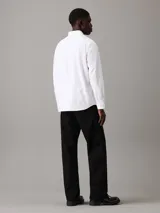Camisa blanca de corte regular, confeccionada en tejido oxford de algodón y elastano. Tiene mangas largas con puños abotonados, cierre frontal con botones y cuello clásico. Incluye etiqueta con el logo de Calvin Klein.