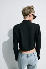 Blazer crop color negro, con cuello subido, manga larga y cierre frontal con botones ocultos. Presenta detalles de alamares bordados a contraste en color dorado en el frente y en los puños.