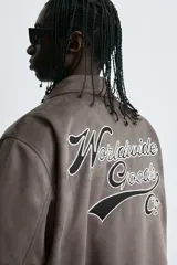 Cazadora estilo varsity de tejido efecto ante en color beige. Presenta cuello solapa, cierre frontal con botones a presión y puños y ruedo con acabado en rib a rayas negras. Incluye parches bordados decorativos en el pecho y la manga, además de bolsillos laterales de doble vivo.
