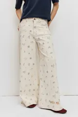 Pantalón de jean wide leg color beige con pequeños bordados y estampados coloridos dispersos, de tiro medio y corte amplio en las piernas.