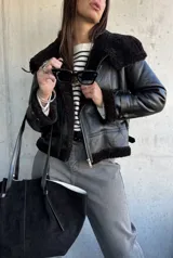 Campera tipo gamulán de cuero sintético negro con interior y detalles de cuello y puños en piel sintética negra. Presenta cierre frontal, correas ajustables en los puños y diseño de corte clásico.