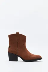 Bota corta estilo western de gamuza color marrón, con taco cuadrado negro y puntera estilizada. Presenta un detalle de estrella aplicada en el lateral y tirador en la parte superior.