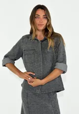 Chaqueta color gris con textura de líneas onduladas, cuello camisero, cierre frontal con botones, mangas 3/4 con puños enrollados, dos bolsillos de parche en el pecho y corte recto.