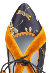Zapato tipo blucher de raso negro con bordado de libélulas en color ocre. Presenta detalles en terciopelo ocre en los laterales y el talón, con un borde festoneado y costuras en zigzag. Se ajusta con cordones de algodón rematados con borlas de ante.