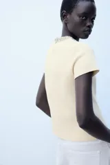 Jersey de punto color amarillo claro, con cuello redondo adornado con aplicación de joyas a contraste, manga corta y cierre en la espalda con abertura y botón.