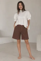 Blusa blanca de lino, con cuello alto, mangas cortas abullonadas con puños con volados y diseño amplio con frunce bajo el busto.