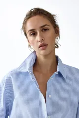 Camisa de corte relajado con diseño a rayas finas en tonos celeste y blanco, cuello clásico, mangas largas con puños anchos abotonados y bajo redondeado.
