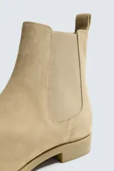 Bota estilo Chelsea de piel con acabado serraje color beige, con elásticos laterales para facilitar el calce, horma redonda y suela a tono con ligero tacón.