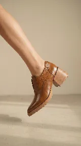 Zapato tipo slip-on color marrón, confeccionado en cuero natural con combinación de texturas lisas y grabadas. Presenta un diseño de punta almendrada, taco cuadrado de madera y suela dentada.