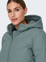 Campera acolchada de corte puffer con capucha, cierre frontal de cremallera y mangas largas con hombros caídos. Presenta un diseño repelente al agua con puños internos elásticos para mayor protección contra el viento.