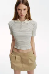 Remera polo de punto acanalado, color crema con finas rayas horizontales negras. Es de manga corta, corte entallado y cuello clásico con tapeta de botones.