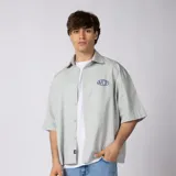 Camisa de manga corta color gris claro, de corte boxy y ajuste ancho, con cuello camisero, cierre frontal de botones y bordado pequeño en el pecho.