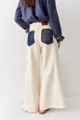 Jean de corte wide leg en color crudo, con detalle de franjas laterales tejidas en crochet azul y verde. Presenta costuras en contraste y ruedo deshilachado.