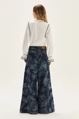 Blusa blanca de algodón con cuerpo de jersey y mangas de popelina. Presenta detalles de punto smock en puños y cuello, adornados con delicados bordados en azul y dorado.