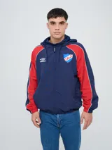 Campera deportiva Umbro del Club Nacional de Football, modelo Venti, para hombre. Diseño en color blanco con mangas azules y detalles de líneas rojas. Cuenta con capucha con cordón ajustable, cierre frontal y bolsillos. Presenta el logo de Umbro y el escudo del club bordados en el pecho.