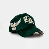 Gorra tipo baseball color verde, con bordado frontal de las letras 'LA' en tipografía gótica y una paloma blanca en el lateral. Cuenta con cierre ajustable en la parte posterior.